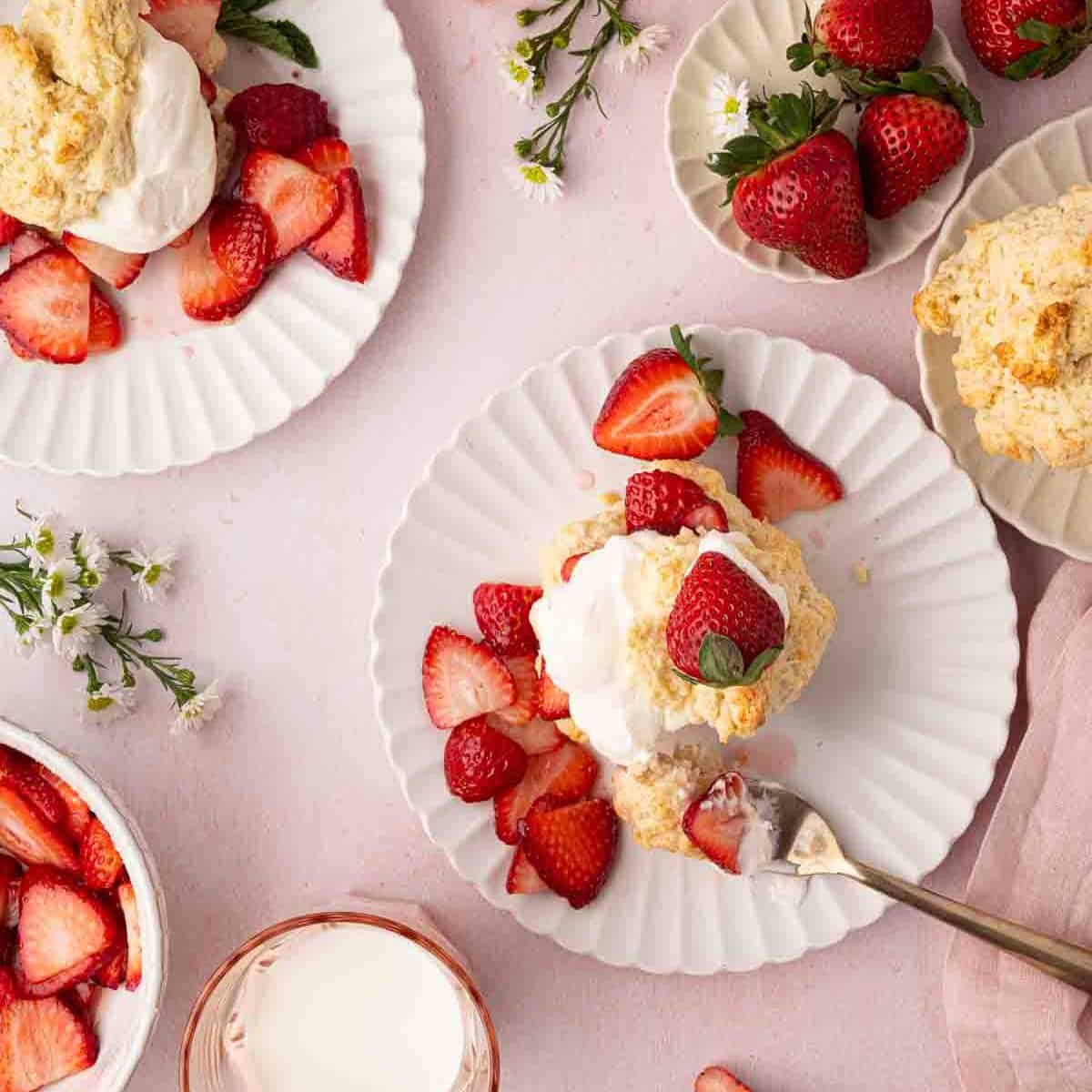 Plates with strawberry shortcakes topped with whipped cream and fresh strawberries, surrounded by whole strawberries, sliced strawberries, flowers, and a glass of milk on a pink surface.