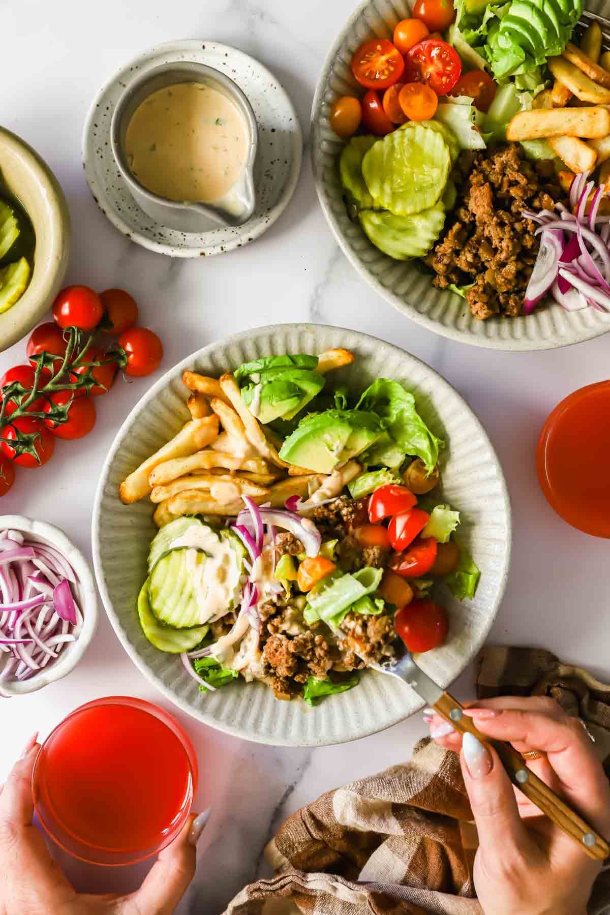 A plate with salad greens, sliced pickles, cherry tomatoes, fries, ground meat, and dressing sits on a table with a drink, sauce, and additional vegetables nearby.