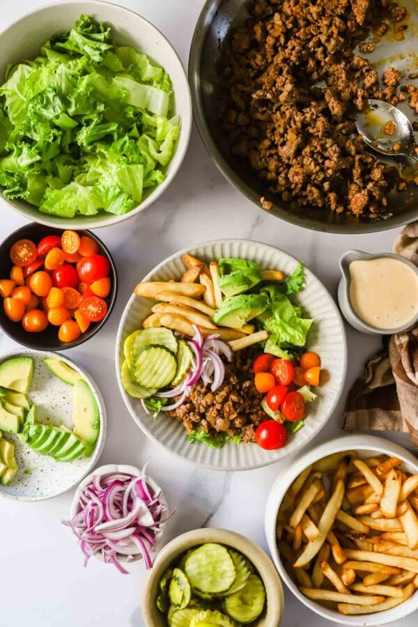 A variety of ingredients are displayed on a table, including lettuce, ground meat, tomatoes, avocado, pickles, red onions, French fries, and a plate with a mixed salad and fries.
