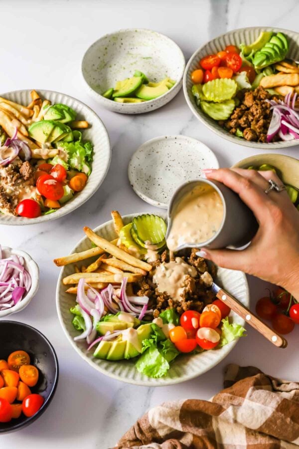 A hand pours dressing onto a bowl filled with seasoned ground meat, fries, cherry tomatoes, sliced avocado, pickles, and red onions; other similar bowls are on the table.