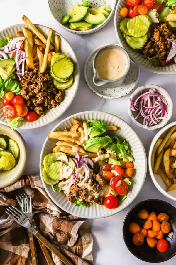 Overhead view of plates with lettuce, ground meat, cherry tomatoes, pickles, fries, sliced red onion, and creamy dressing, along with bowls of avocado and extra toppings on the side.