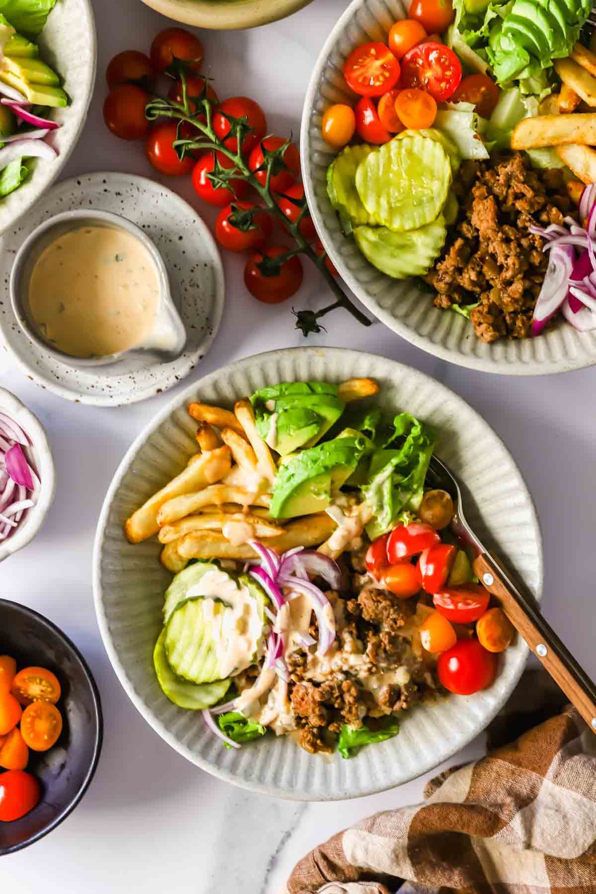 Two bowls filled with ground meat, lettuce, tomato, sliced cucumber, red onion, and French fries, served with a small dish of creamy sauce and fresh cherry tomatoes on the side.