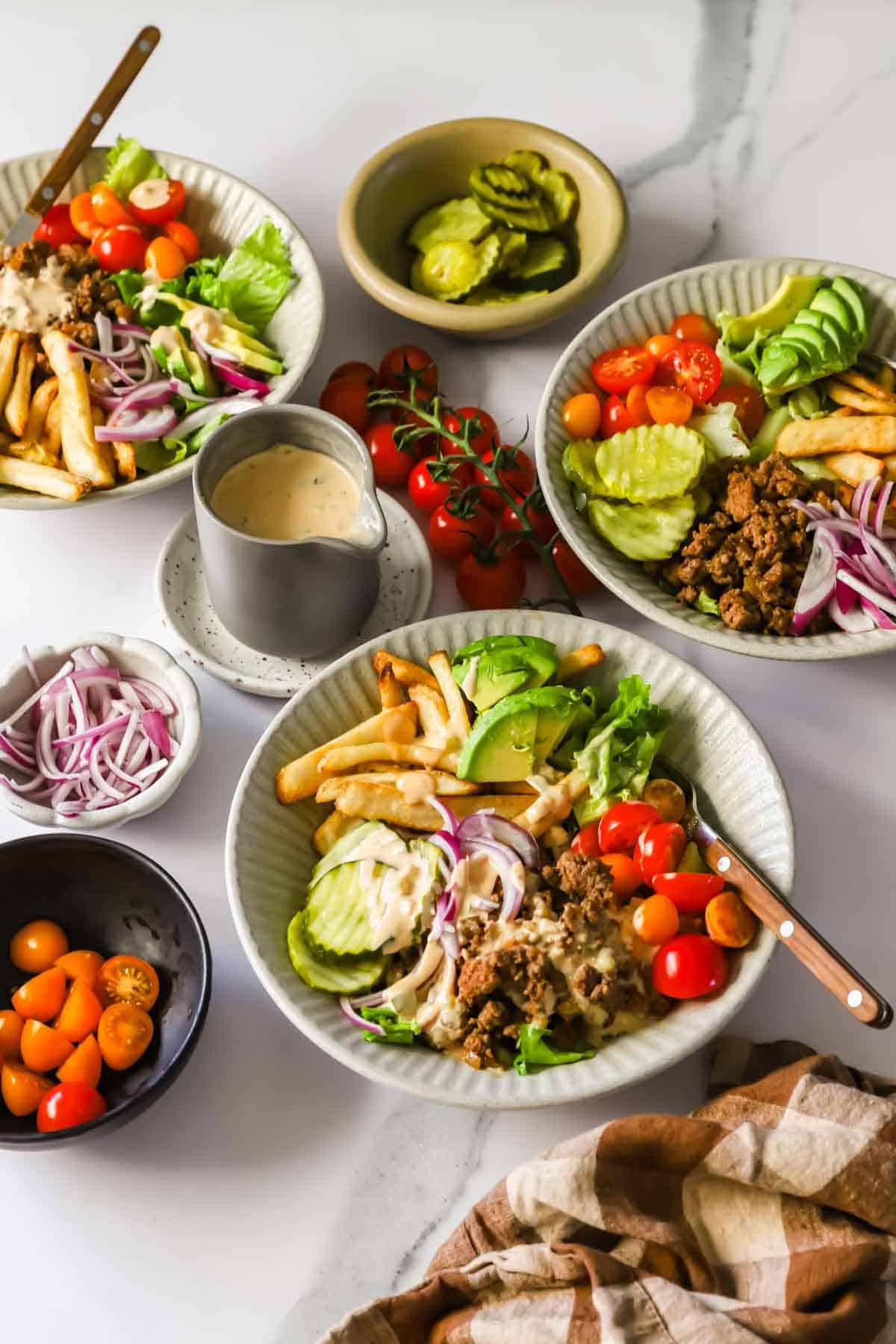 Three bowls of salad with ground beef, French fries, pickles, cherry tomatoes, red onions, lettuce, and avocado, surrounded by extra toppings and dressing on a marble surface.
