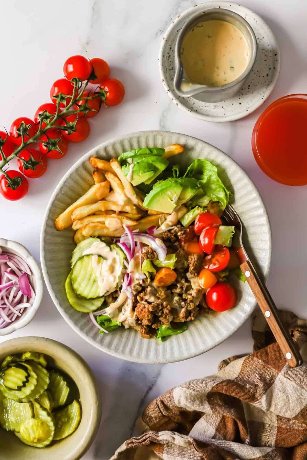 A plate with salad, french fries, ground meat, sliced cucumbers, tomatoes, red onions, and dressing. Surrounding are a bowl of pickles, cherry tomatoes, dressing, and a glass of juice.