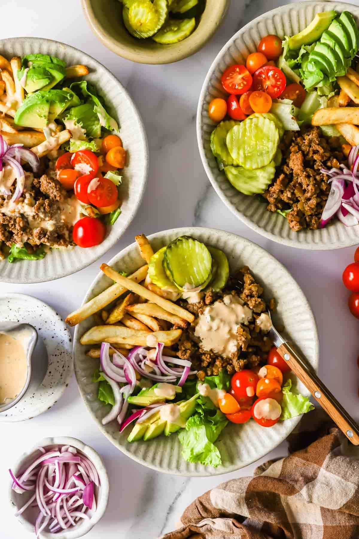Three plates with ground beef, lettuce, tomatoes, pickles, red onions, French fries, and dressing. Small bowls with pickles, extra red onions, and a pitcher of dressing are nearby.