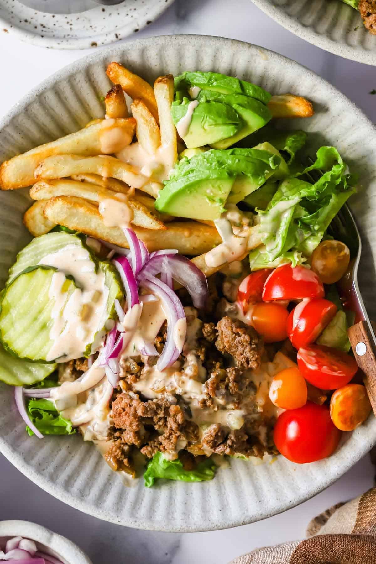 A bowl filled with French fries, sliced avocado, lettuce, pickles, red onion, cherry tomatoes, ground beef, and a drizzle of creamy dressing.