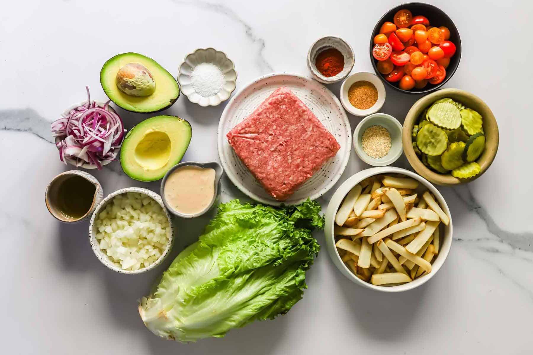 Ingredients for a lettuce wrap burger arranged on a white surface, including ground meat, lettuce, fries, pickles, tomatoes, avocado, onion, seasonings, and sauce.