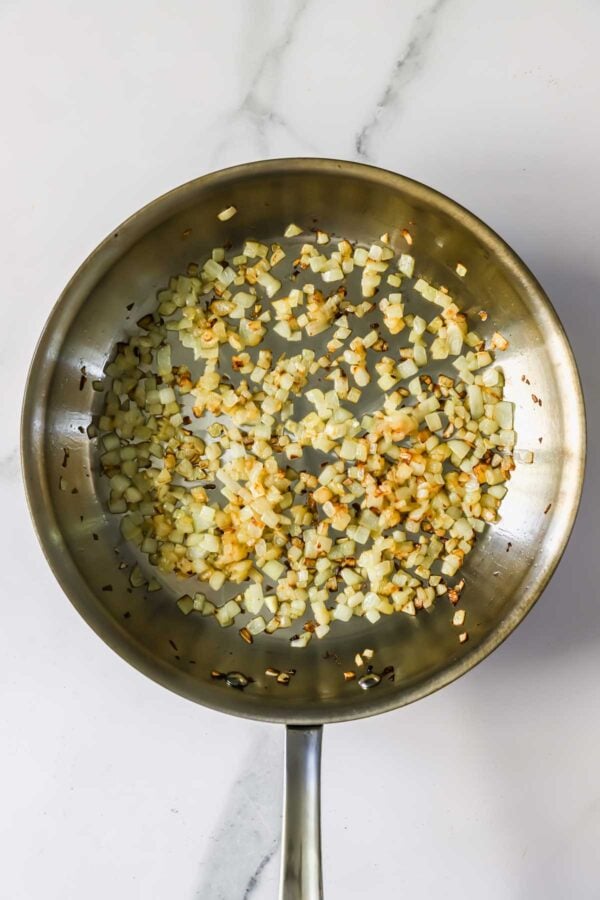 A stainless steel pan with diced onions being saut&eacute;ed, some pieces turning golden brown, on a white marble surface.