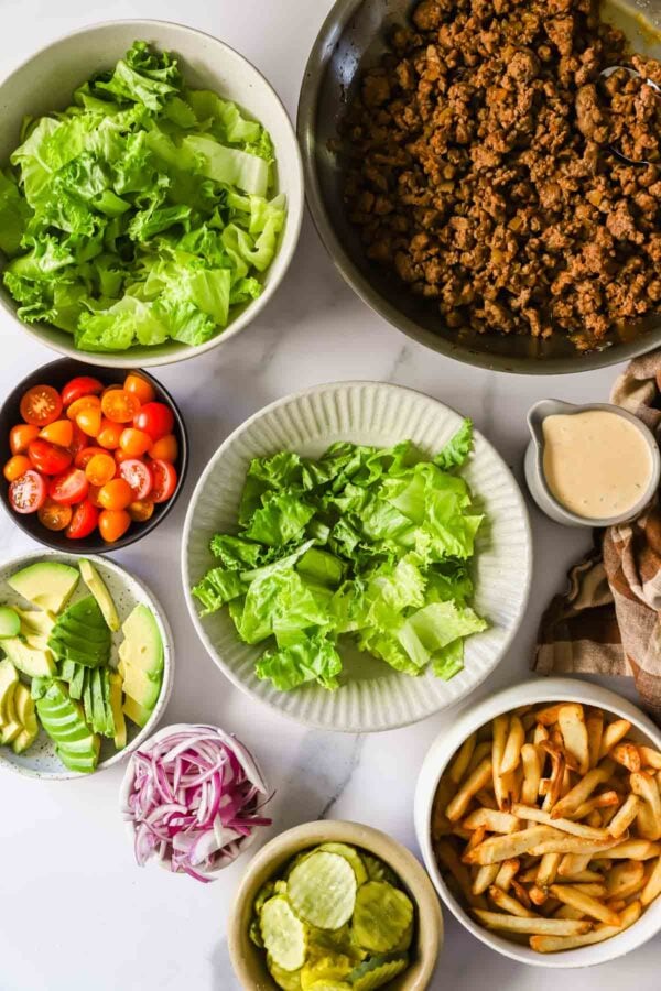 Overhead view of burger bowl ingredients: cooked ground beef, lettuce, cherry tomatoes, avocado, red onion, pickles, French fries, and a small pitcher of sauce.