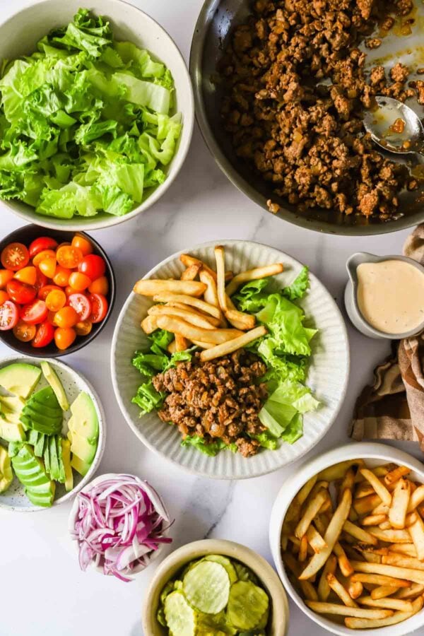 A flat lay of bowls with lettuce, cherry tomatoes, avocado, red onion, pickles, fries, cooked ground meat, and salad dressing, with one plate assembled with fries, lettuce, and meat.