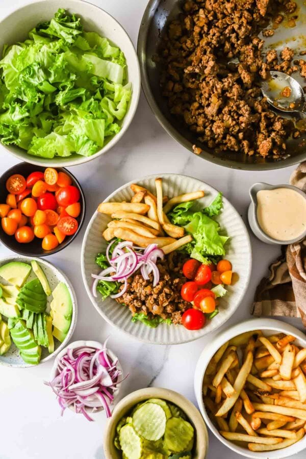 An overhead view of various burger bowl ingredients, including ground beef, lettuce, cherry tomatoes, avocado, red onion, pickles, fries, and a small pitcher of sauce.