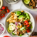 A plate with ground beef, French fries, lettuce, cucumber, cherry tomatoes, and red onion slices, with a sauce and drink nearby.