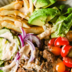 A burger bowl with ground beef, sliced avocado, tomato halves, lettuce, fries, red onion, and creamy sauce on a plate. Text reads "Best Burger Bowl Recipe.