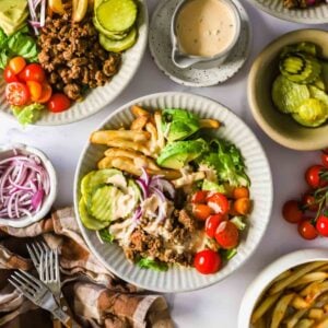 A plate with ground beef, lettuce, tomato, avocado, pickles, French fries, and onions, topped with a light sauce. Extra toppings, fries, and sauce are on the side.