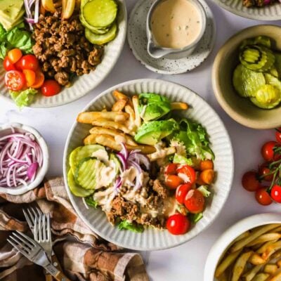 A plate with ground beef, lettuce, tomato, avocado, pickles, French fries, and onions, topped with a light sauce. Extra toppings, fries, and sauce are on the side.