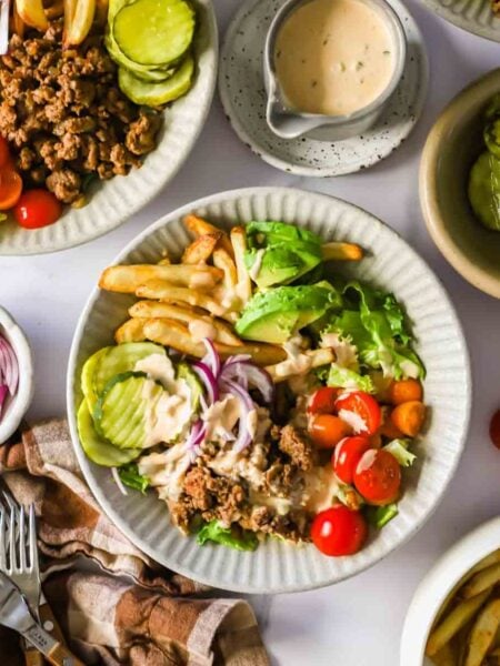 A plate with ground beef, lettuce, tomato, avocado, pickles, French fries, and onions, topped with a light sauce. Extra toppings, fries, and sauce are on the side.