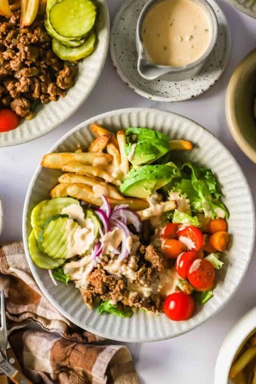 A plate with ground beef, lettuce, tomato, avocado, pickles, French fries, and onions, topped with a light sauce. Extra toppings, fries, and sauce are on the side.