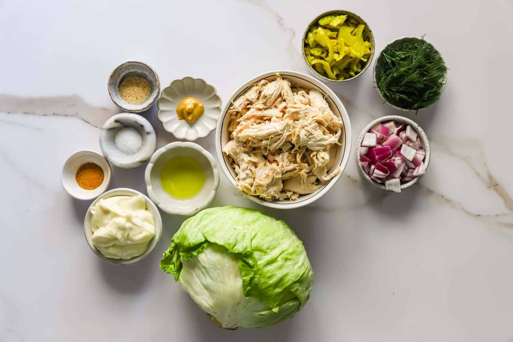 Overhead view of shredded chicken, iceberg lettuce, chopped red onions, dill, pepper rings, mayonnaise, olive oil, mustard, and seasonings on a white surface.