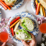 A person holds a chicken salad lettuce wrap surrounded by plates with wraps, carrot and celery sticks, and glasses of pink beverage on a table.