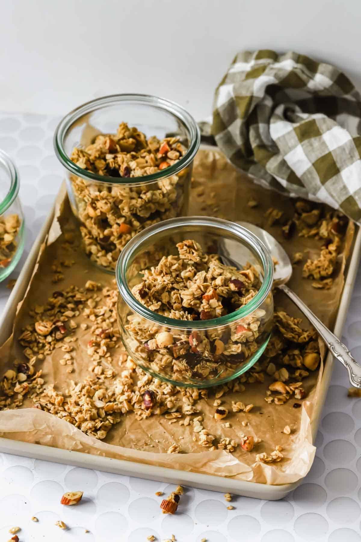Three glass jars filled with maple granola sit on a parchment-lined baking tray, with a spoon and a checkered cloth nearby.