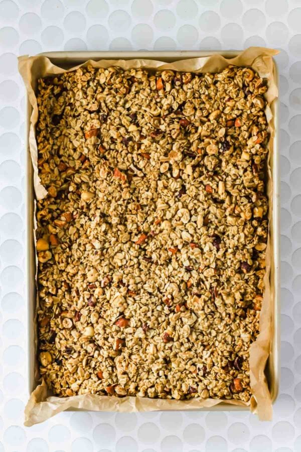 A baking sheet lined with parchment paper holding a single, even layer of homemade granola with visible oats and mixed nuts.