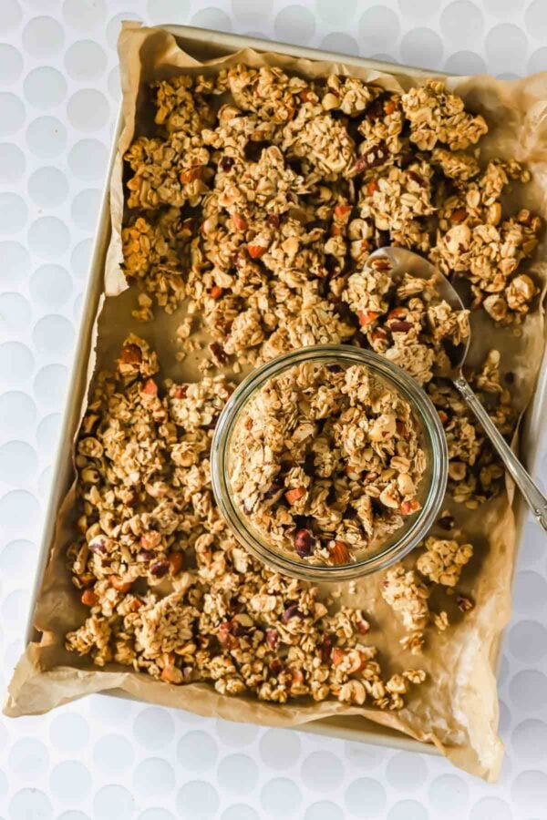 A baking tray lined with parchment paper holds clusters of homemade granola, with some placed in a glass jar and a spoon resting beside it.