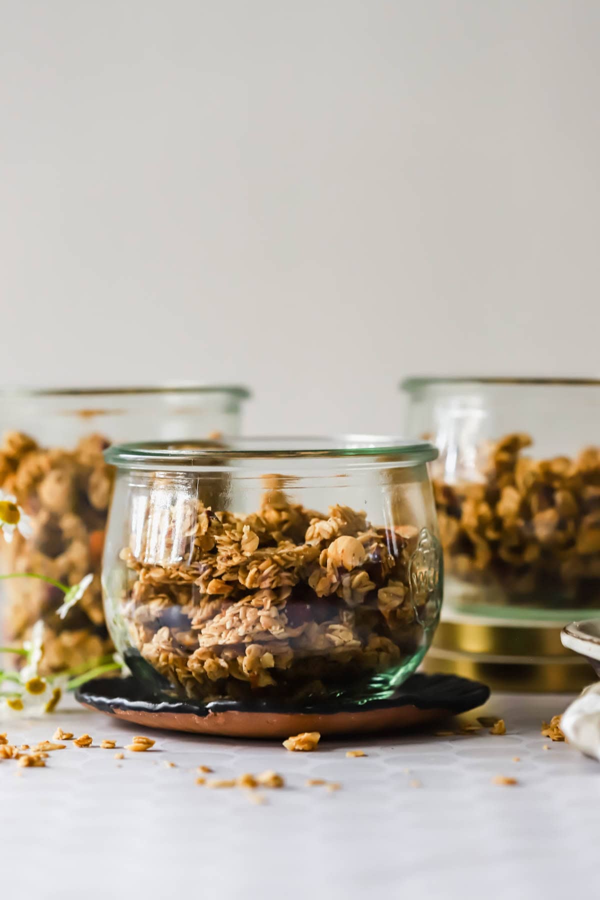 Three glass jars filled with granola clusters are displayed on a white surface with a neutral background.