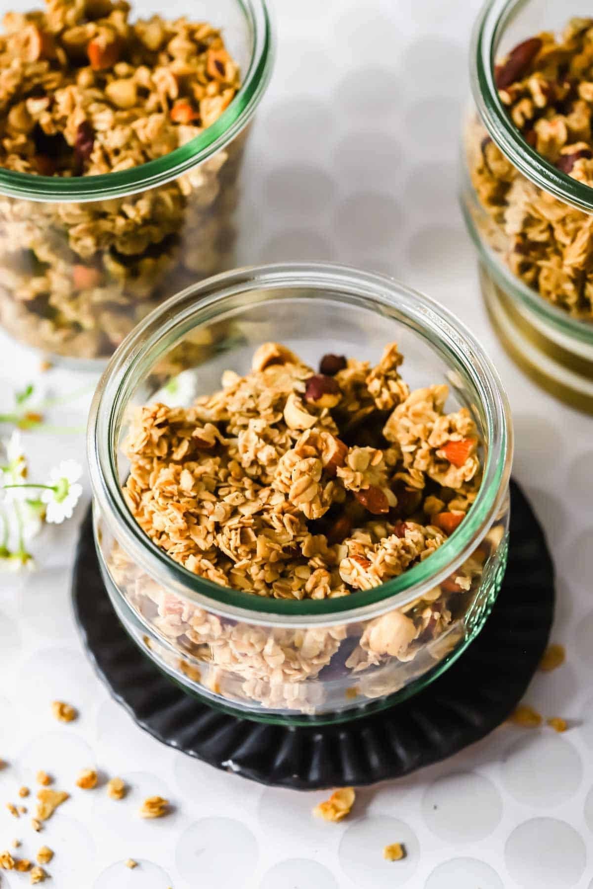 Glass jars filled with homemade granola containing oats and nuts, placed on a white surface with scattered granola pieces.