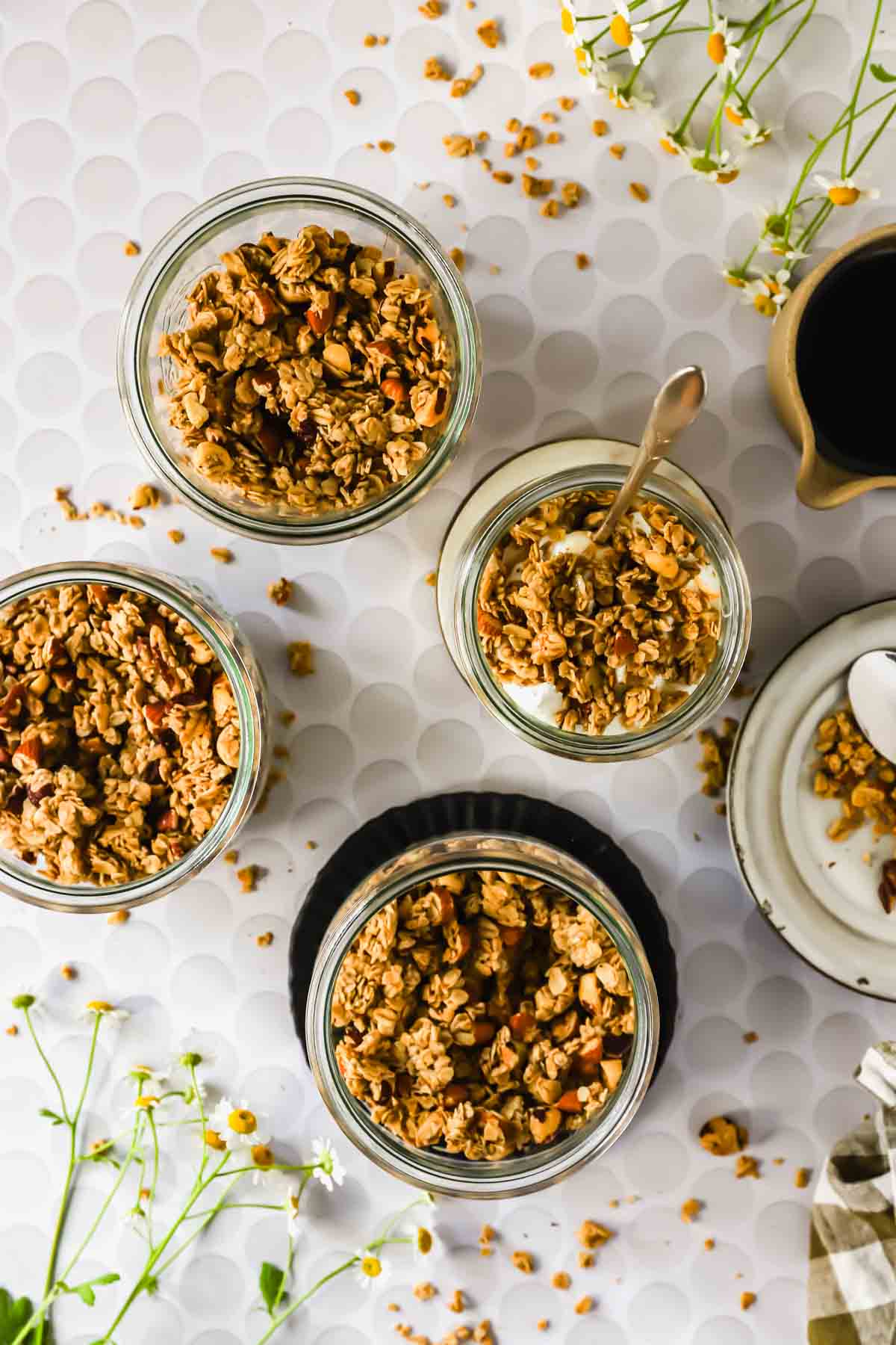 Four glass jars filled with granola, one with yogurt and a spoon, are arranged on a white patterned surface with scattered flowers and a small pitcher of dark liquid.