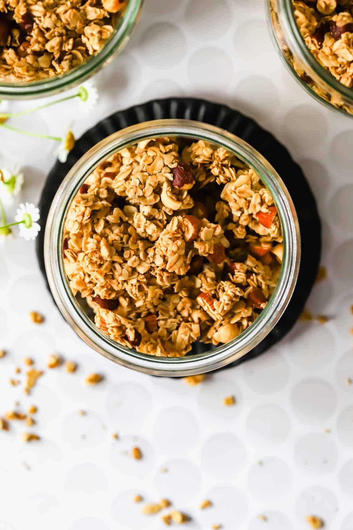 A glass jar filled with granola, viewed from above, with some granola pieces scattered on a white surface nearby.