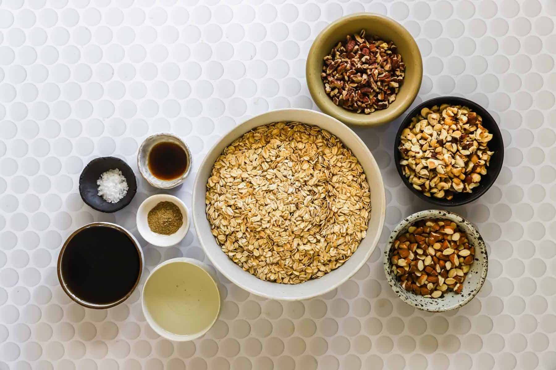 A large bowl of rolled oats surrounded by small bowls containing chopped nuts, seeds, vanilla extract, oil, syrup, and salt on a white textured surface.