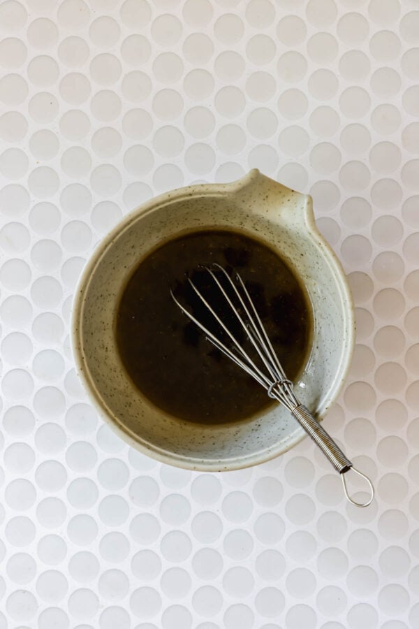 A ceramic bowl with a dark liquid mixture and a metal whisk inside, placed on a white textured circular tile surface.