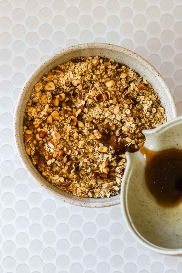 Bowl of oats and chopped nuts with a dark liquid being poured from a small jug, set on a white textured surface.
