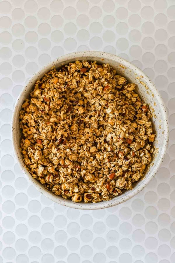 A white bowl filled with granola containing oats and chopped nuts, placed on a white textured surface.
