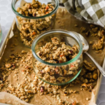 Two glass jars filled with homemade maple granola sit on a baking sheet lined with parchment paper, with a spoon, scattered granola, and a checked cloth nearby.