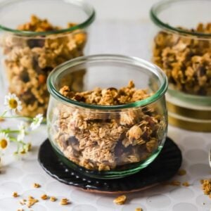 Three glass jars filled with granola are arranged on a table with loose granola pieces and small white flowers nearby.