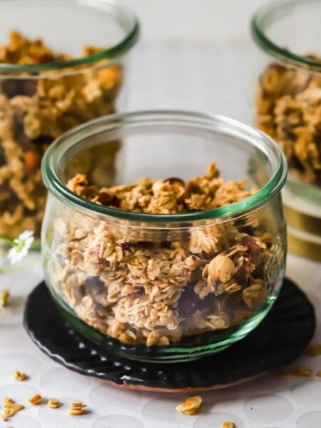 Three glass jars filled with granola are arranged on a table with loose granola pieces and small white flowers nearby.