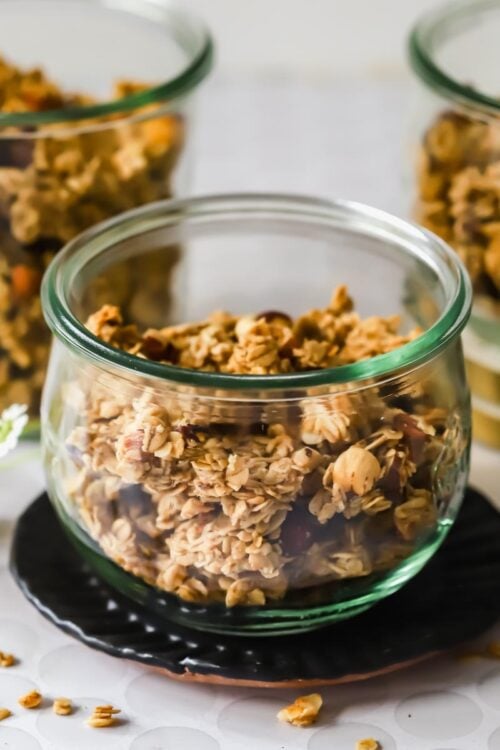 Three glass jars filled with granola are arranged on a table with loose granola pieces and small white flowers nearby.