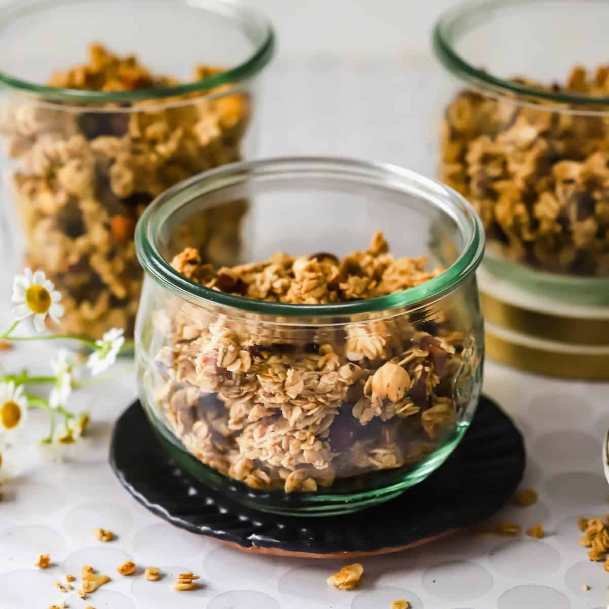 Three glass jars filled with granola are arranged on a table with loose granola pieces and small white flowers nearby.