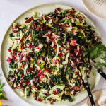 A plate of radicchio fennel salad with chopped greens, nuts, and herbs, served with two black utensils on a white table next to a sliced orange and a cup.