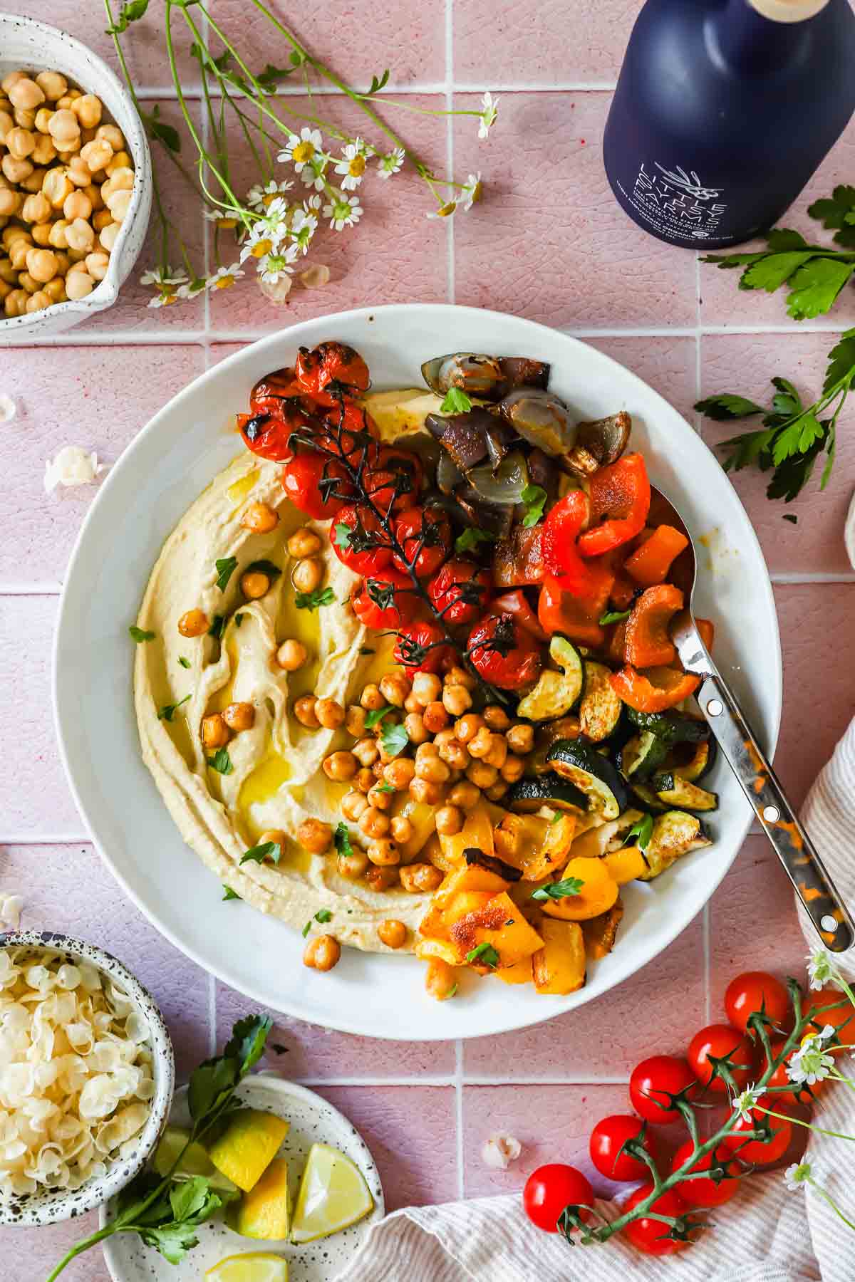 A white plate of hummus topped with roasted cherry tomatoes, zucchini, bell peppers, eggplant, and chickpeas, surrounded by bowls of chickpeas, lemon wedges, and fresh herbs on a pink tiled surface.