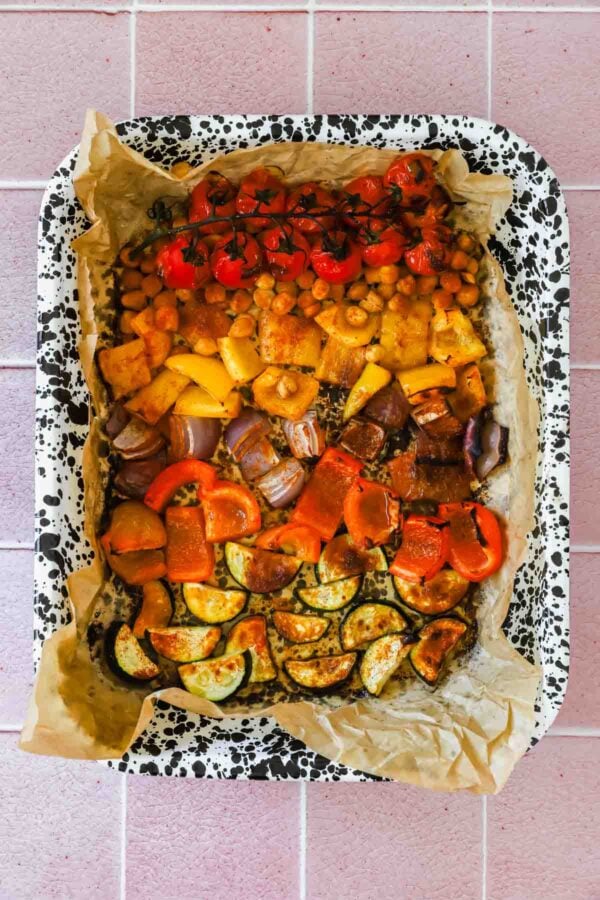A baking tray lined with parchment paper holds an assortment of roasted vegetables, including zucchini, bell peppers, tomatoes, red onion, and chickpeas.