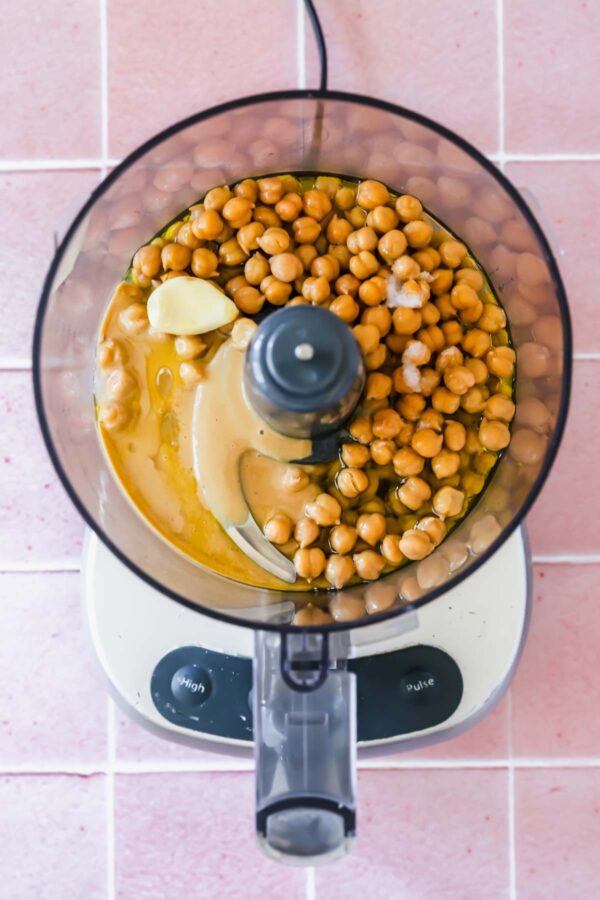 Overhead view of a food processor containing chickpeas, tahini, olive oil, garlic, and other ingredients on a pink tiled surface.