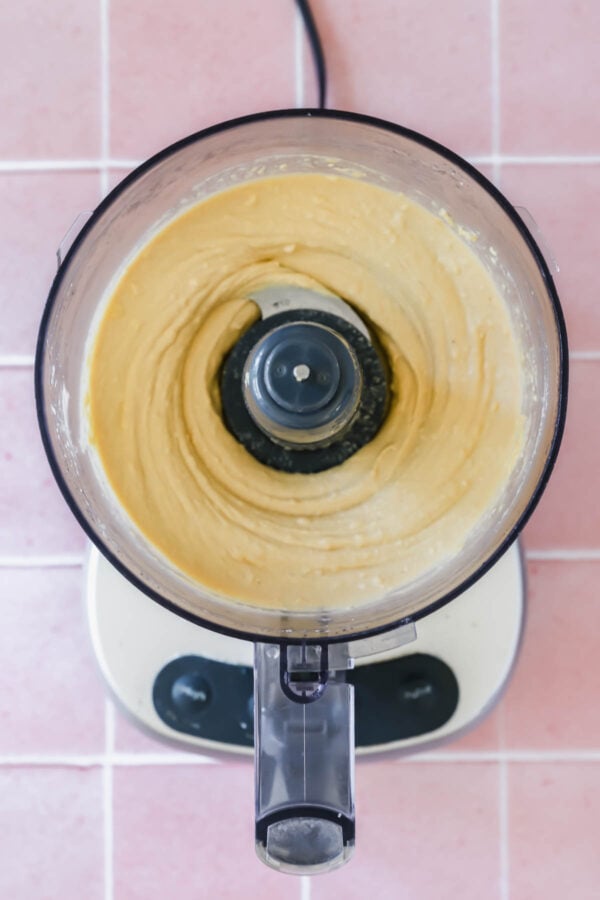 Overhead view of a food processor containing a smooth, creamy mixture on a pink tiled surface.