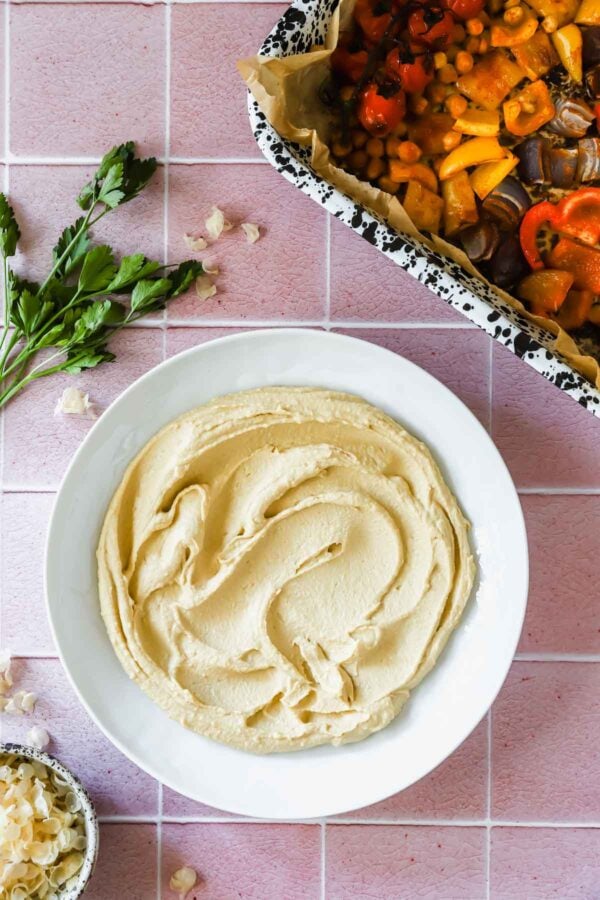 A plate of hummus on a pink tiled surface next to a tray of roasted vegetables and fresh parsley.