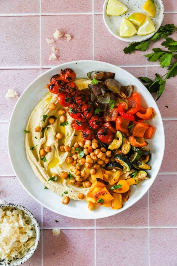 A white bowl of hummus topped with roasted tomatoes, chickpeas, red onion, bell peppers, and zucchini, placed on a pink tiled surface with lemon wedges and parsley nearby.