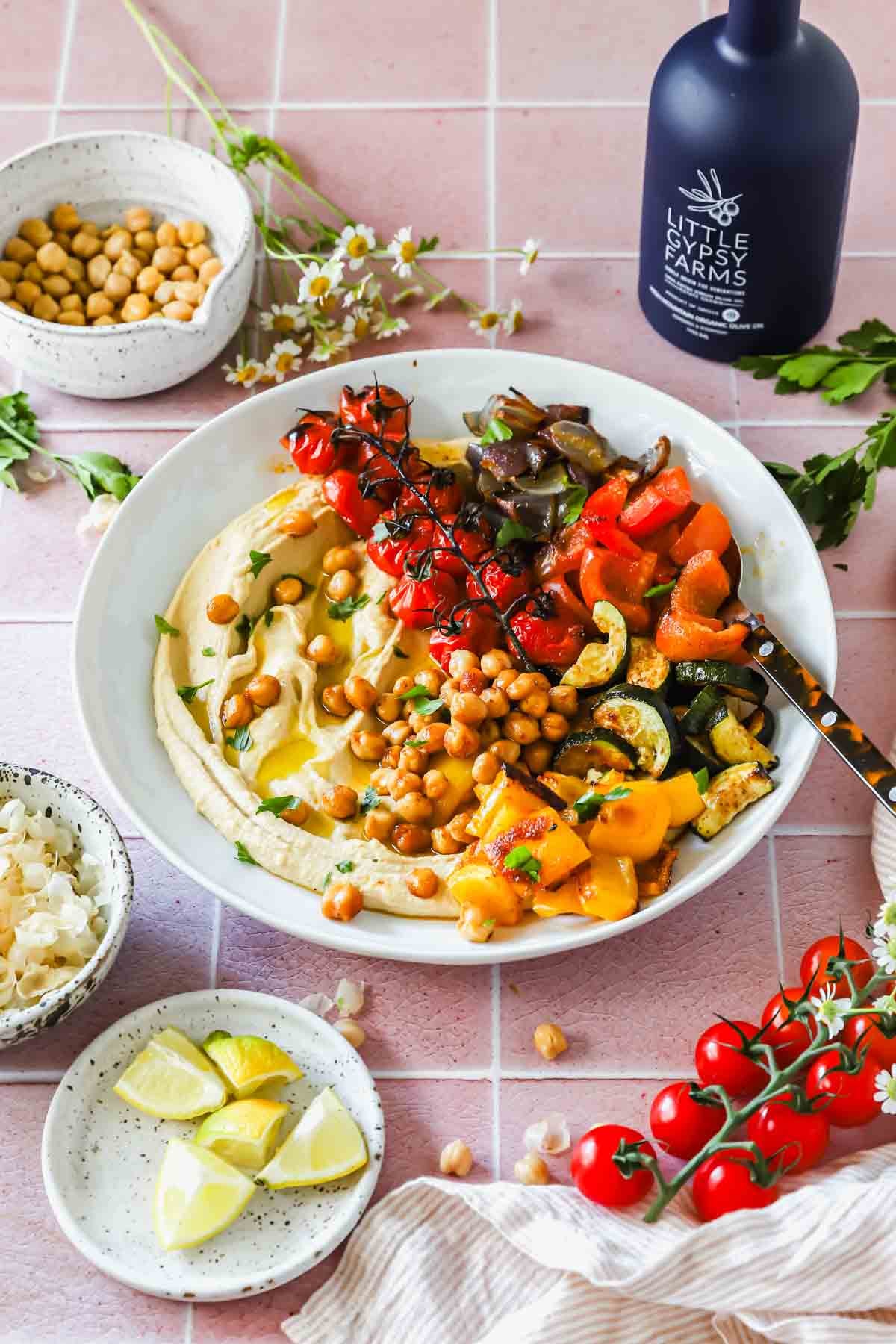 A bowl of hummus topped with roasted vegetables and chickpeas, surrounded by lime wedges, cherry tomatoes, chickpeas, and a bottle of olive oil on a pink tiled surface.