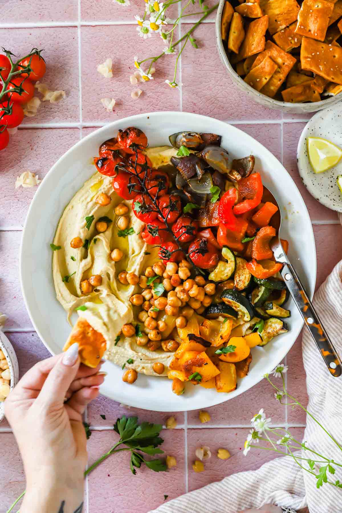A plate of hummus topped with roasted vegetables and chickpeas, served with pita chips. A hand dips a chip into the hummus.