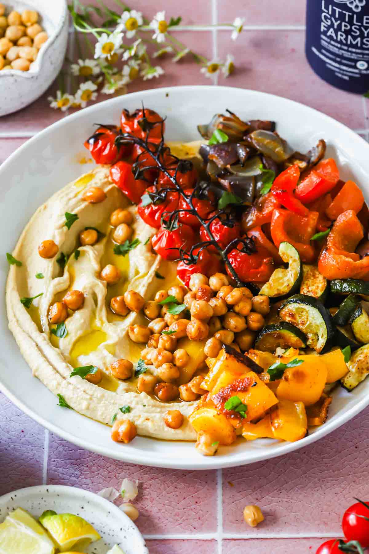 A bowl of hummus topped with roasted chickpeas, cherry tomatoes on the vine, diced red and yellow bell peppers, zucchini, and eggplant, garnished with parsley.