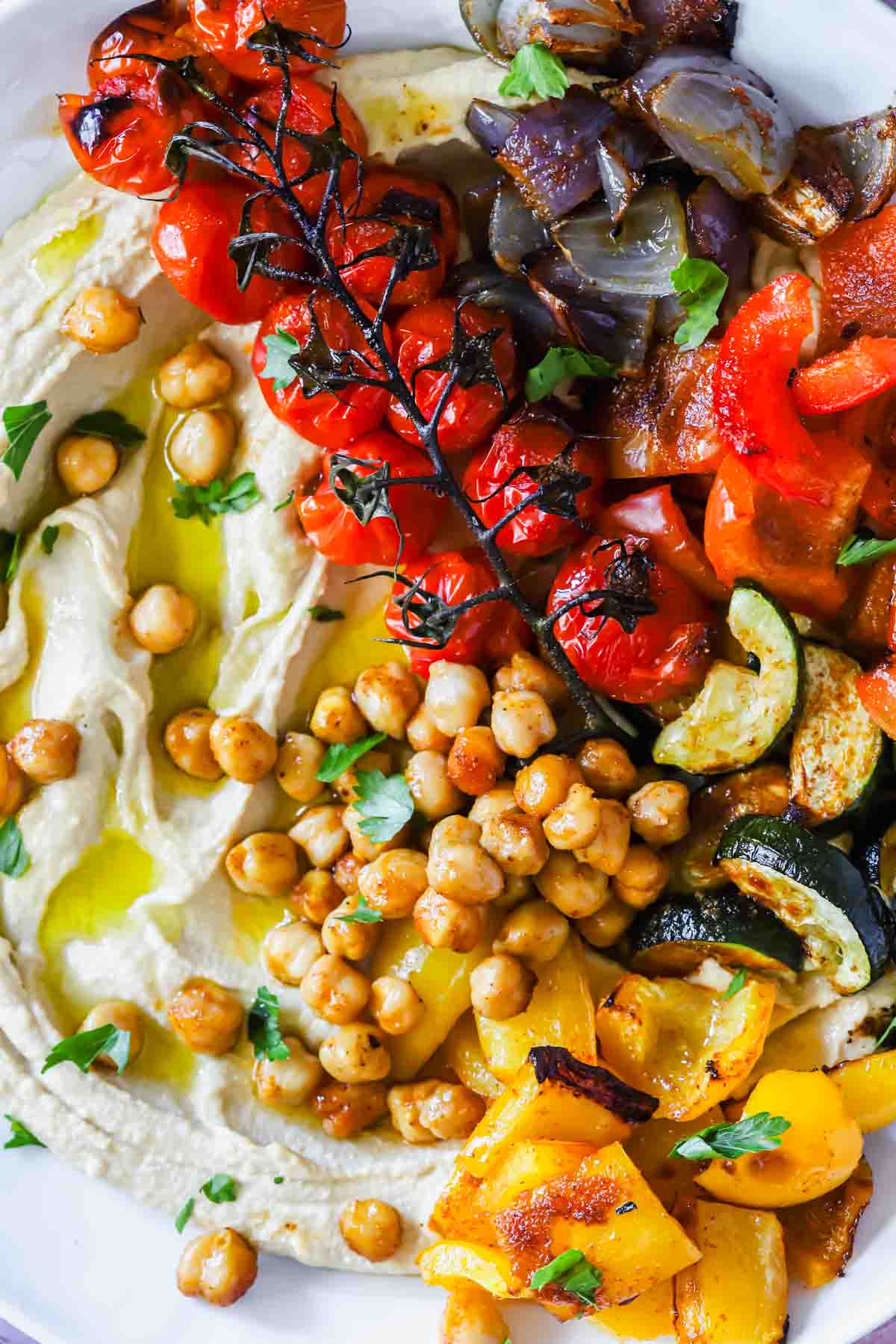 A plate of hummus topped with roasted chickpeas, tomatoes on the vine, and assorted roasted vegetables including peppers, zucchini, and red onion, garnished with fresh herbs.